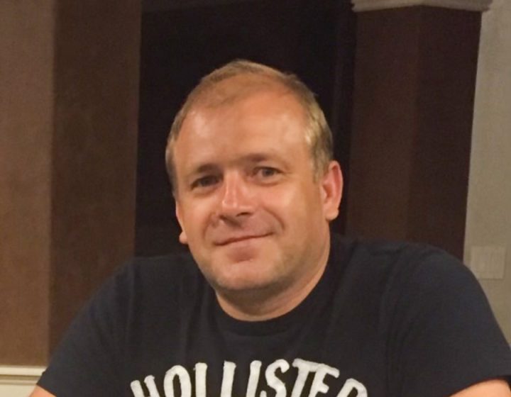 Head-and-shoulders portrait of a smiling male flooring professional wearing a navy Hollister t-shirt, seated indoors with architectural columns and warm ambient lighting in the background.