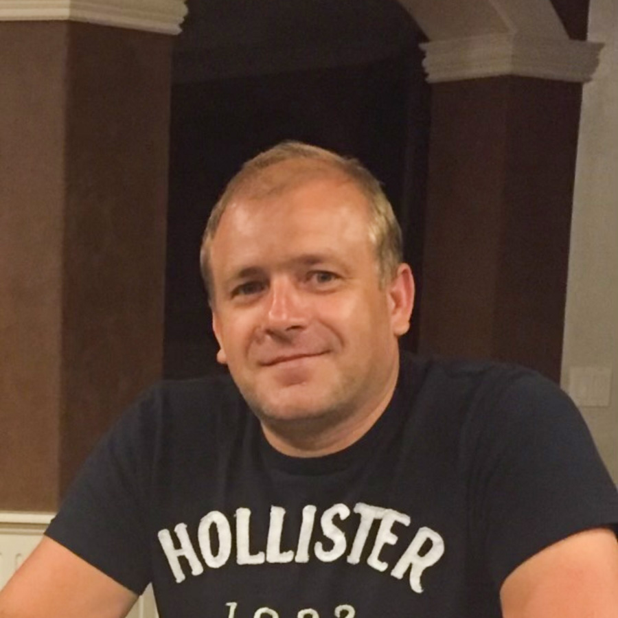 Head-and-shoulders portrait of a smiling male flooring professional wearing a navy Hollister t-shirt, seated indoors with architectural columns and warm ambient lighting in the background.