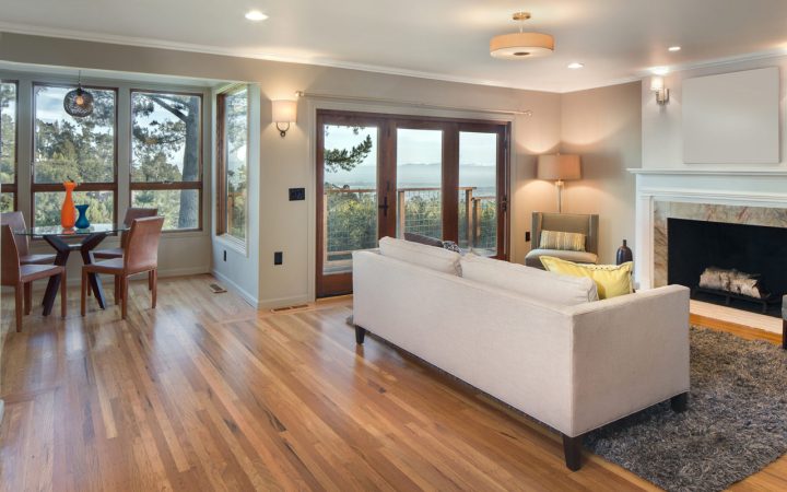 Hero image of a sunlit open-concept living and dining area featuring warm wide-plank oak hardwood flooring, floor-to-ceiling windows with forest and mountain views, neutral furnishings, and a marble fireplace surround.